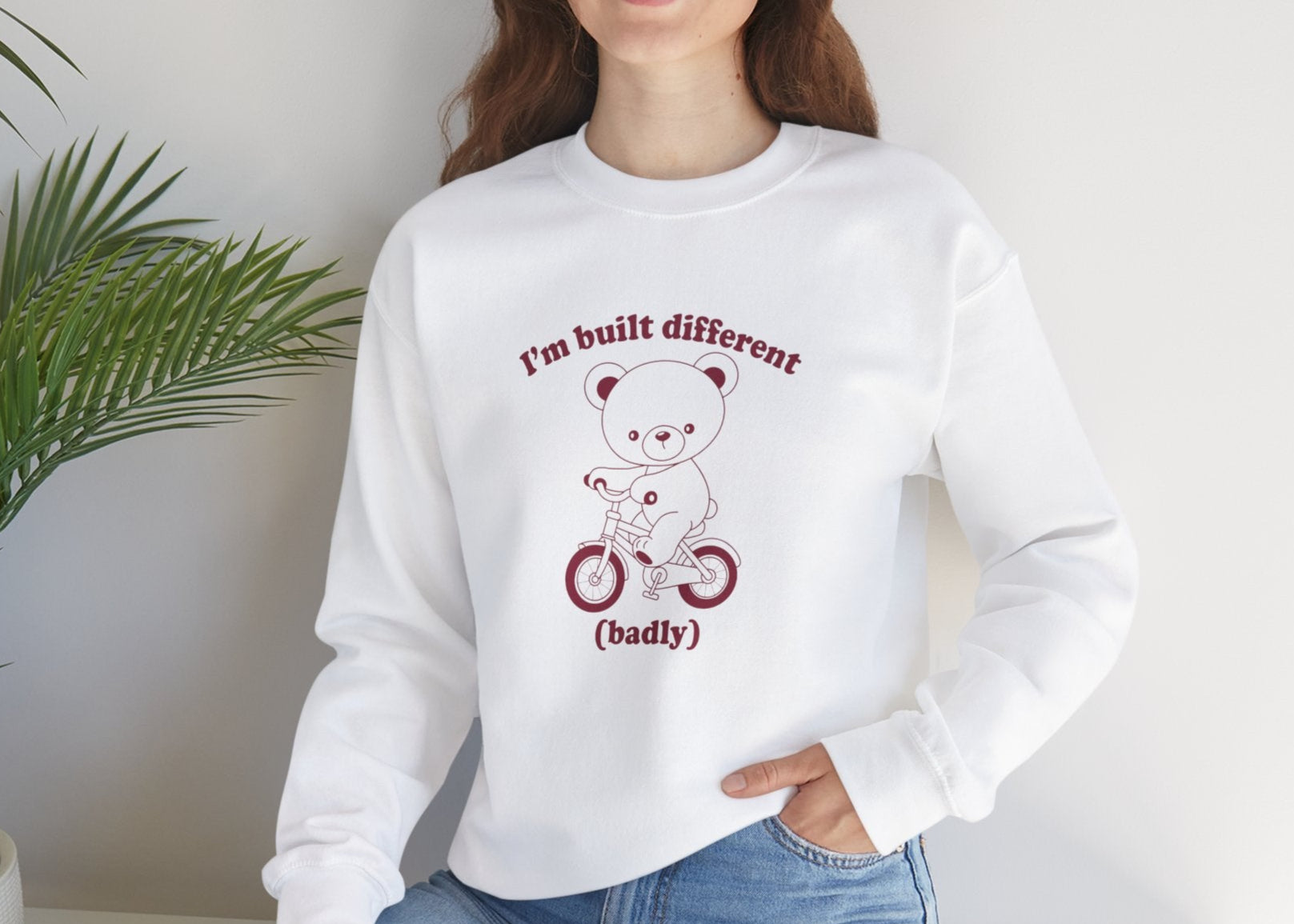 Model seated, wearing a white sweatshirt with a graphic of a bear riding a bike and the text "I'm Built Different (Badly)." Part of the Vivid Divergence Sensory Friendly Unisex Sweater Jumper Range.