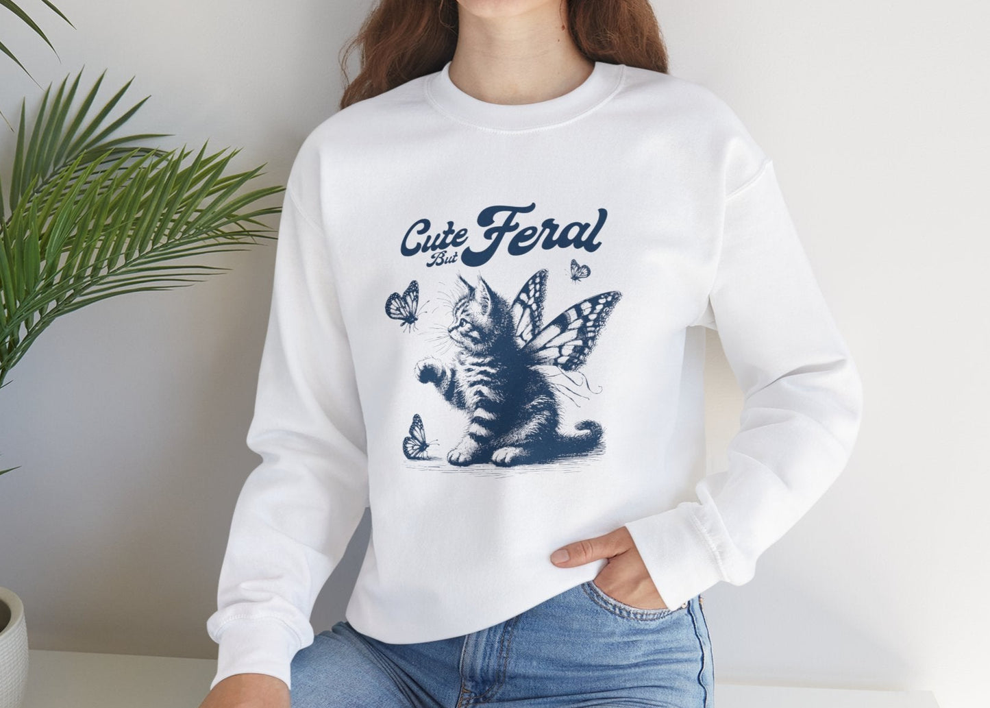 Model seated, wearing a white sweatshirt with a graphic of a cat surrounded by butterflies and the text "Cat Feral." Part of the Vivid Divergence Sensory Friendly Unisex Sweater Jumper Range.