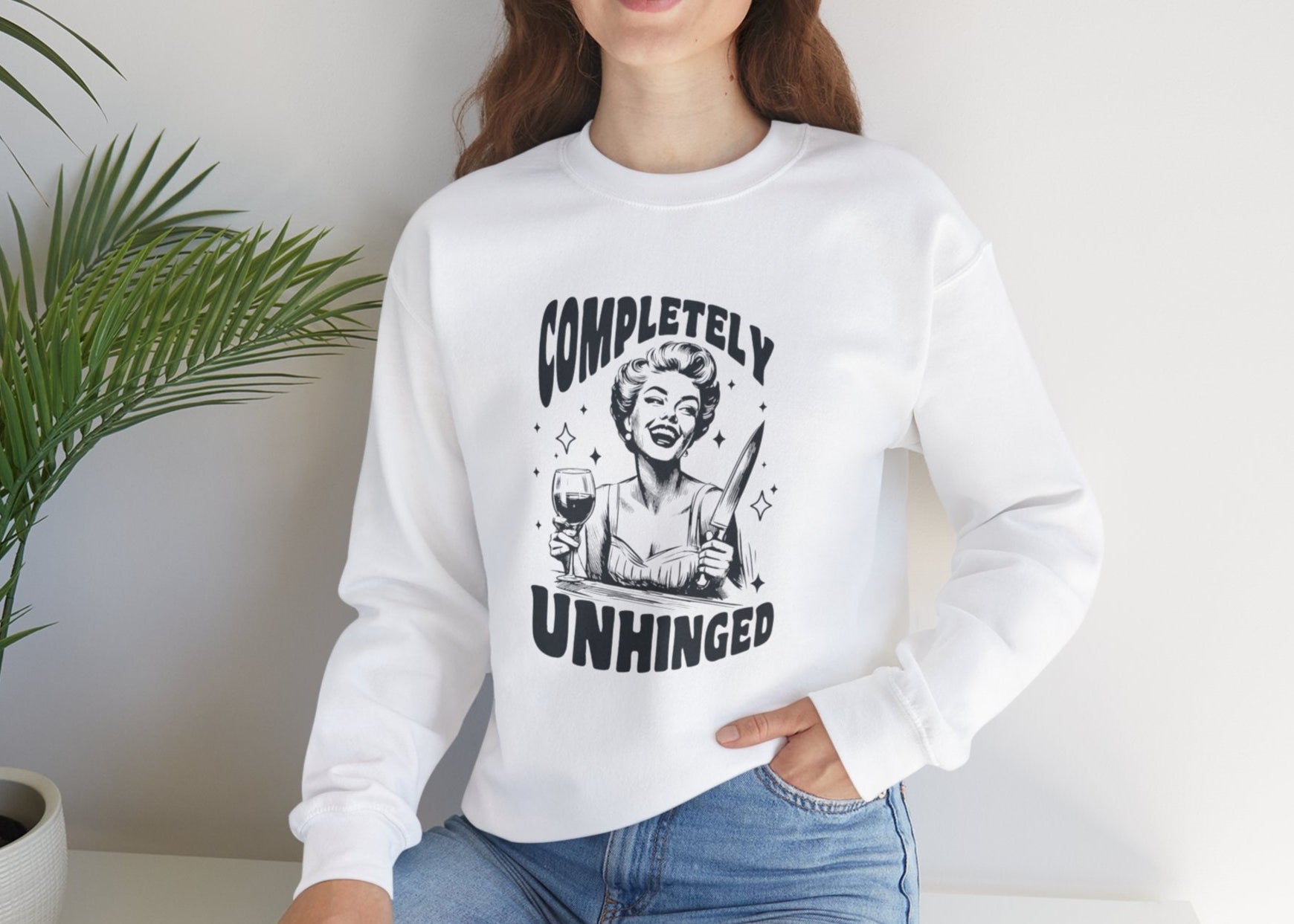 Model seated, wearing a white sweatshirt with a graphic of a woman holding a knife and a glass of wine and the text "Completely Unhinged." Part of the Vivid Divergence Sensory Friendly Unisex Sweater Jumper Range.