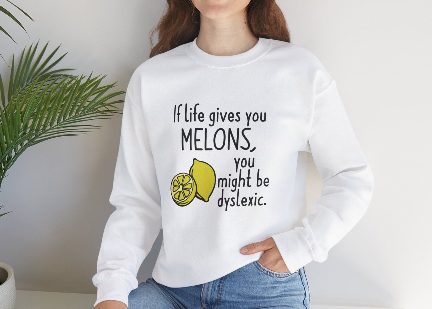 Model seated, wearing a white sweatshirt with an illustration of melons and the text "If Life Gives You Melons, You Might Be Dyslexic." Part of the Vivid Divergence Sensory Friendly Unisex Sweater Jumper Range.