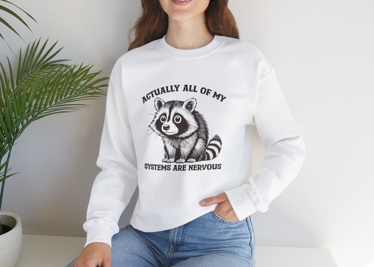 Model seated, wearing a white sweatshirt with a raccoon graphic and the text "Actually All of My Systems Are Nervous." Part of the Vivid Divergence Sensory Friendly Unisex Sweater Jumper Range.