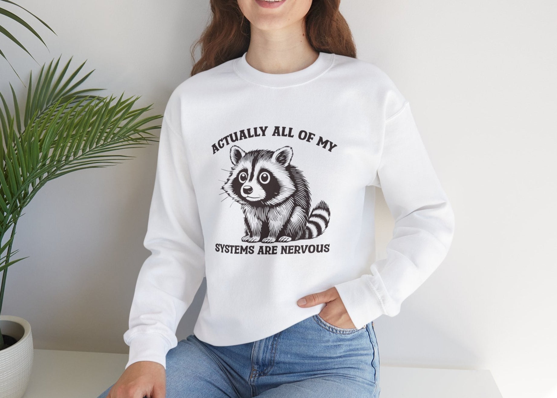 Model seated, wearing a white sweatshirt with a raccoon graphic and the text "Actually All of My Systems Are Nervous." Part of the Vivid Divergence Sensory Friendly Unisex Sweater Jumper Range.