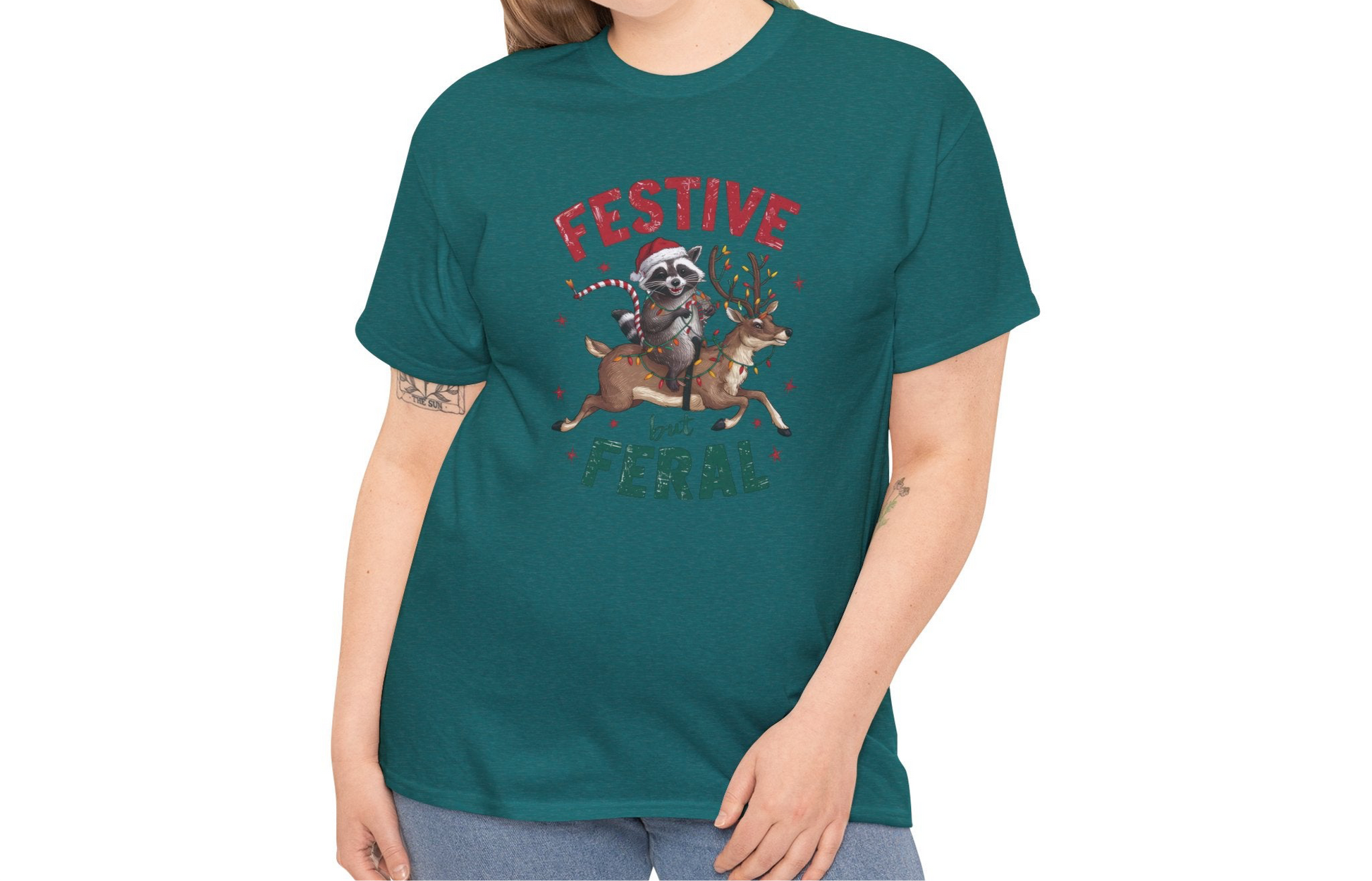 Model wearing a teal T-shirt featuring a festive illustration of a raccoon holding Christmas lights and the text "Festive but Feral," styled casually for the holidays. Part of the Vivid Divergence Sensory Friendly Unisex Tees Range.