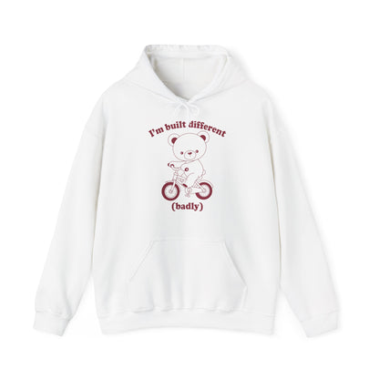 Front view of a white hoodie featuring a cartoon bear riding a small bicycle with the text "I'm Built Different (Badly)." Part of the Vivid Divergence Sensory Friendly Unisex Hoodie Range.
