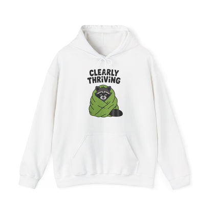 Front view of a white hoodie featuring the text "Clearly Thriving" and a cartoon illustration of a raccoon wrapped in a cozy blanket. Part of the Vivid Divergence Sensory Friendly Unisex Hoodie Range.