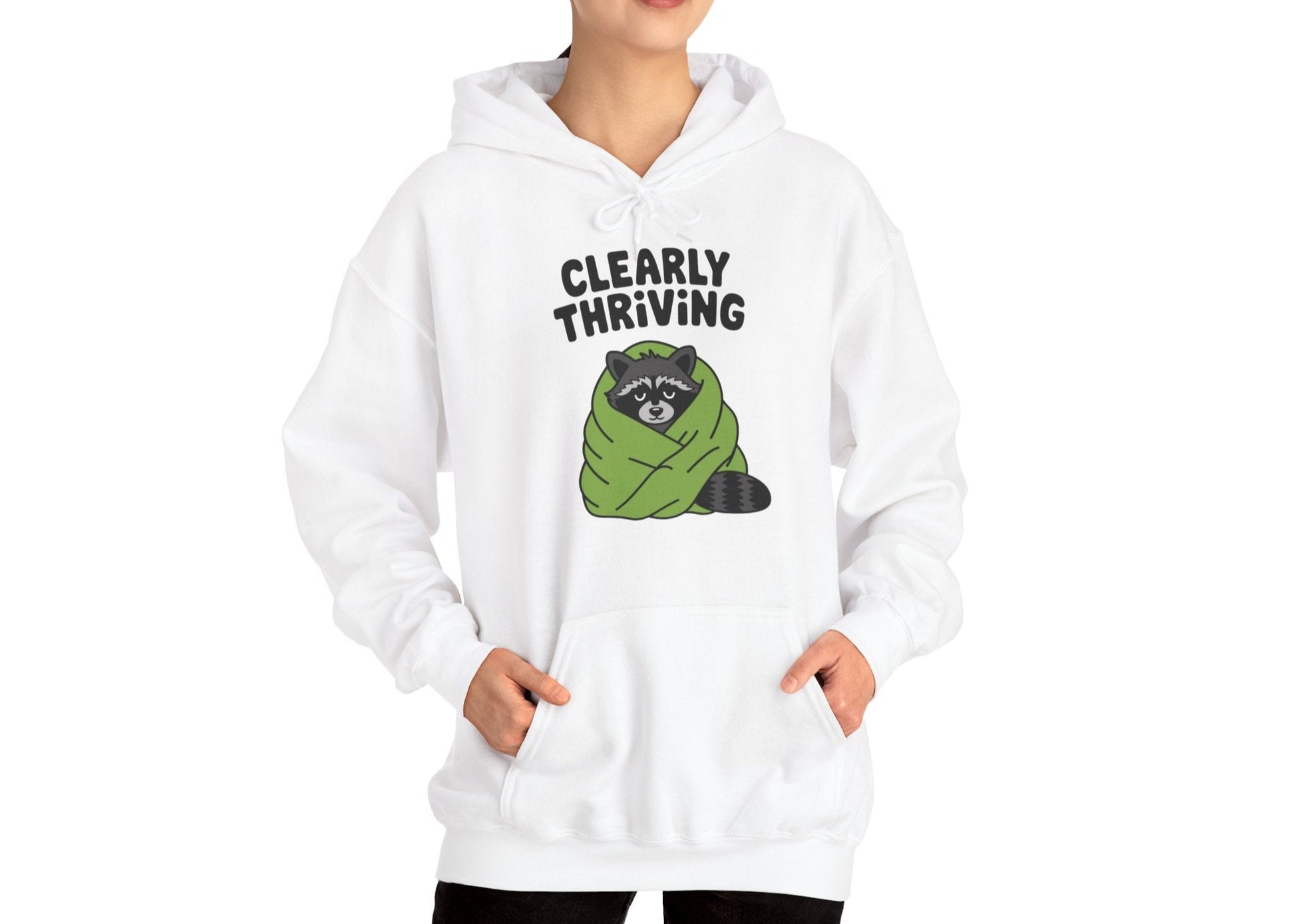 Model wearing a white hoodie featuring the text "Clearly Thriving" and a cartoon illustration of a raccoon wrapped in a cozy blanket. Part of the Vivid Divergence Sensory Friendly Unisex Hoodie Range.