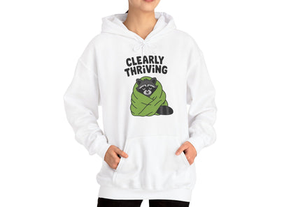 Model wearing a white hoodie featuring the text "Clearly Thriving" and a cartoon illustration of a raccoon wrapped in a cozy blanket. Part of the Vivid Divergence Sensory Friendly Unisex Hoodie Range.