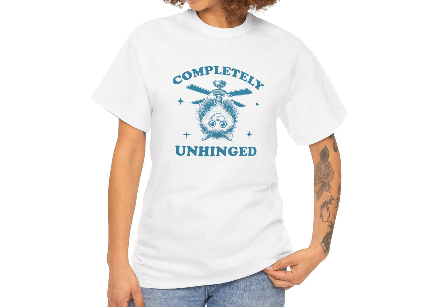 Model with curly hair wearing a white T-shirt featuring "Completely Unhinged" text with a cat hanging upside down from a ceiling fan in blue-tone design. Part of the Vivid Divergence Sensory Friendly Unisex Tees Range.