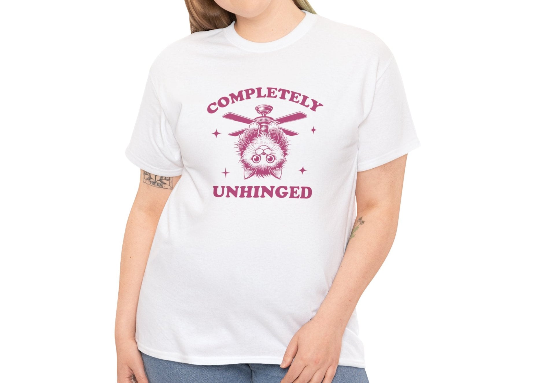 Model wearing a white T-shirt featuring "Completely Unhinged" text with a cat hanging upside down from a ceiling fan in red-tone design. Part of the Vivid Divergence Sensory Friendly Unisex Tees Range.