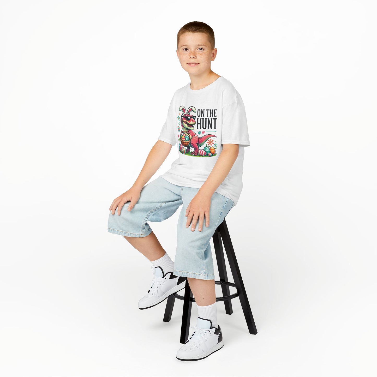 Child wearing a white Easter T-shirt with a dinosaur, "On the Hunt" text, and colorful eggs, sitting on a stool. A cool and festive Easter look. Hop into the season in style and shop Easter shirts online today!