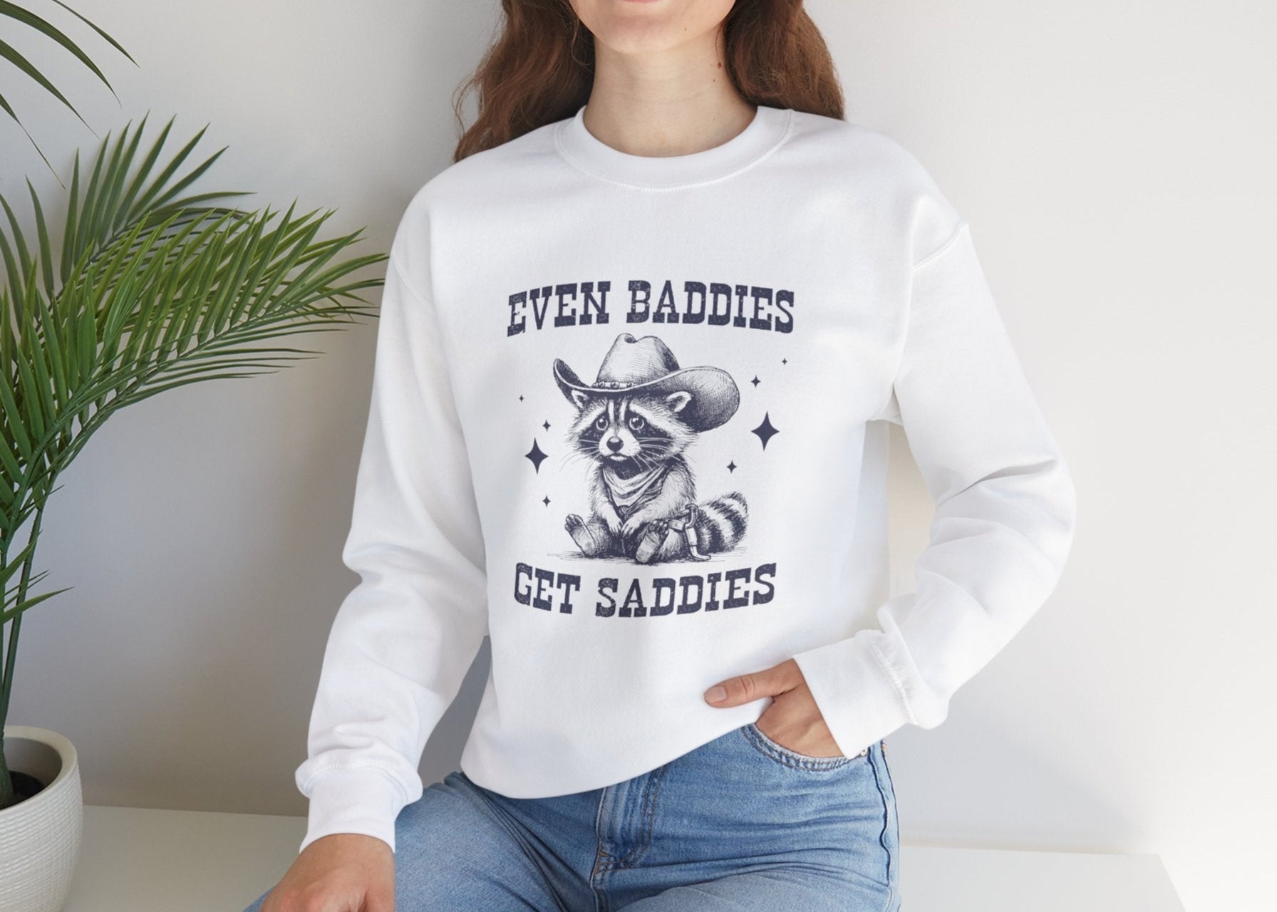 Model wearing a white sweater jumper with the text "Even Baddies Get Saddies" and a cartoon raccoon illustration. Part of the Vivid Divergence Sensory Friendly Unisex Sweater Jumper Range.