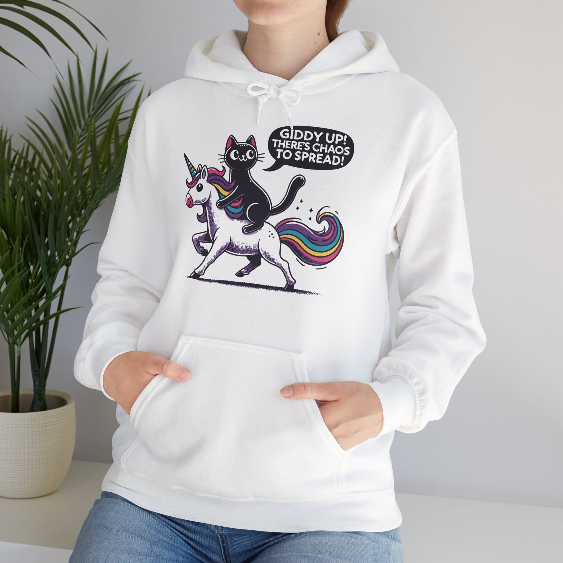 Model sitting while wearing a white hoodie featuring the text "Giddy Up, There’s Chaos to Spread" and a cartoon illustration of a cat riding a unicorn. Part of the Vivid Divergence Sensory Friendly Unisex Hoodie Range.