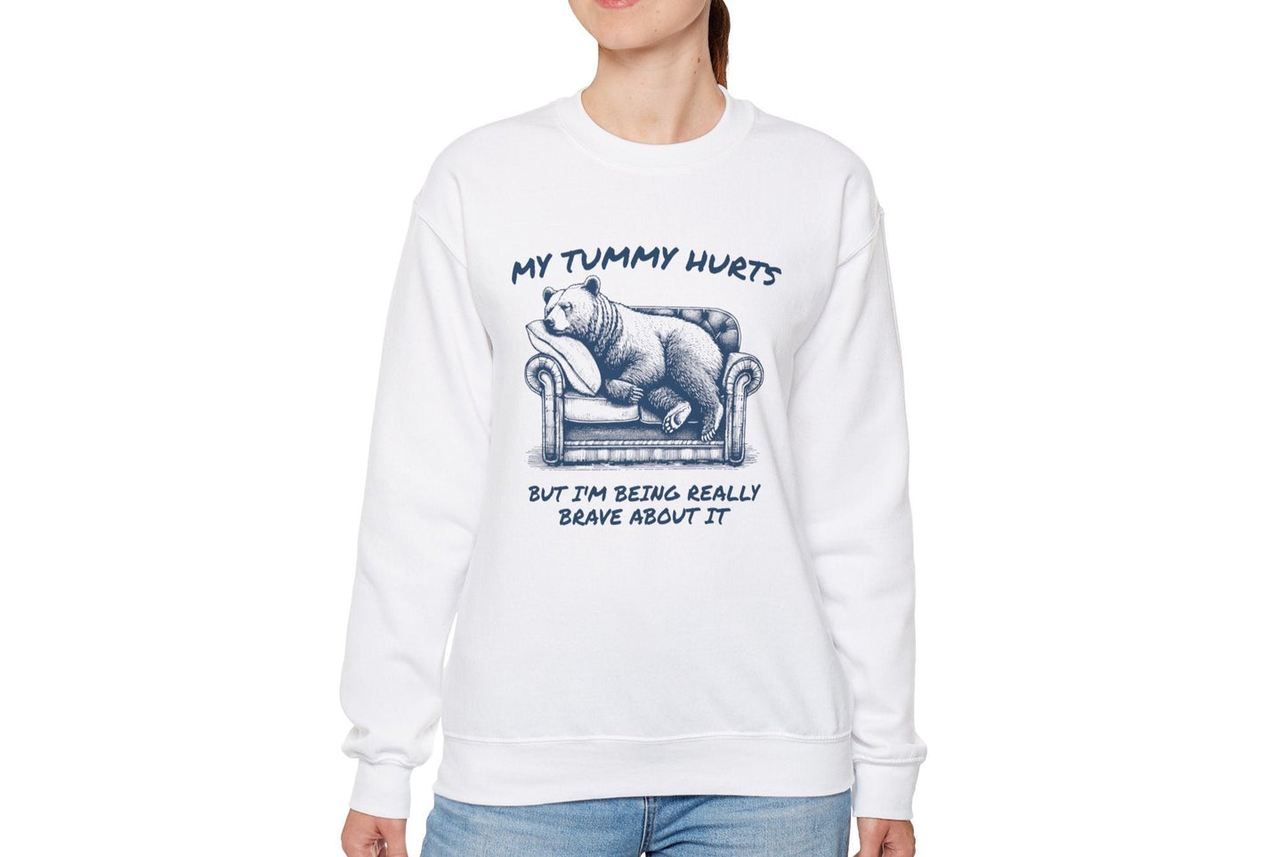Model wearing a white sweater jumper with the text "My Tummy Hurts But I’m Being Really Brave About It" and a cartoon illustration of a bear holding its stomach. Part of the Vivid Divergence Sensory Friendly Unisex Sweater Jumper Range.