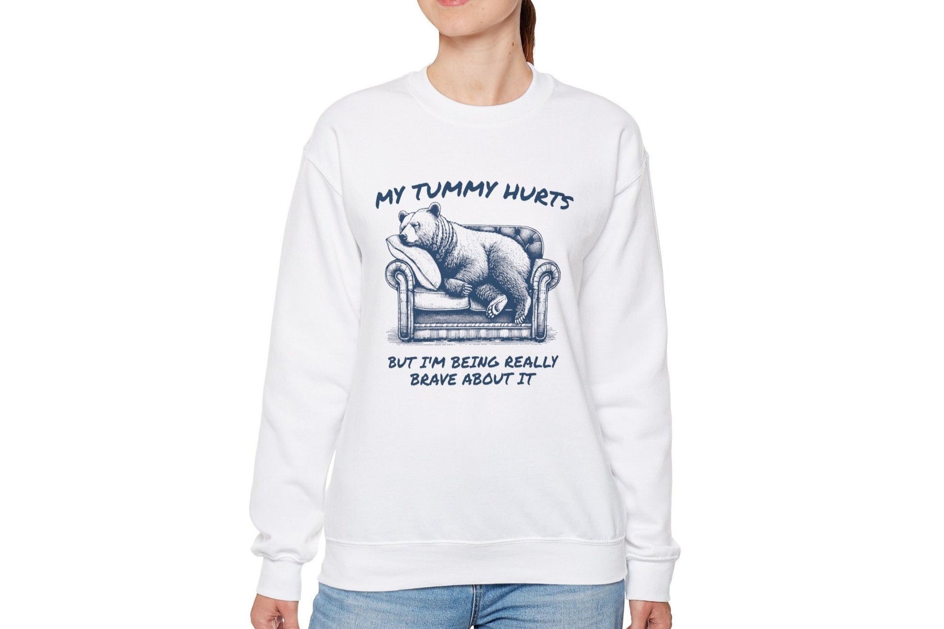Model wearing a white sweater jumper with the text "My Tummy Hurts But I’m Being Really Brave About It" and a cartoon illustration of a bear holding its stomach. Part of the Vivid Divergence Sensory Friendly Unisex Sweater Jumper Range.