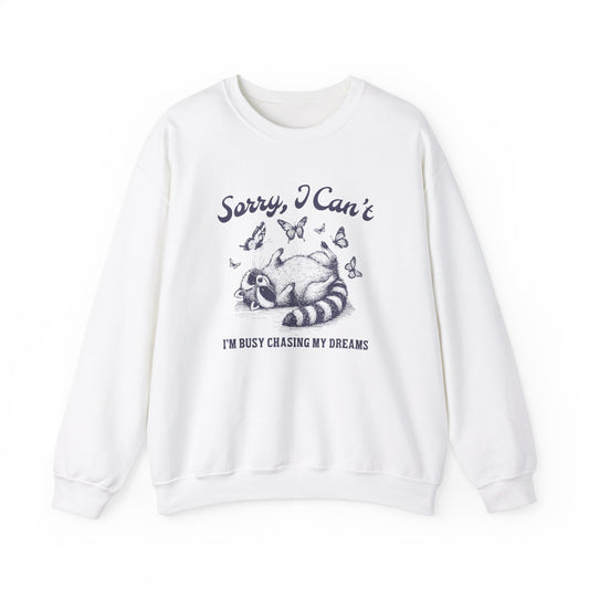 Front view of a white sweater jumper with the text "Sorry, I Can't. I'm Busy Chasing My Dreams" and a cartoon raccoon illustration sleeping on a crescent moon. Part of the Vivid Divergence Sensory Friendly Unisex Sweater Jumper Range.