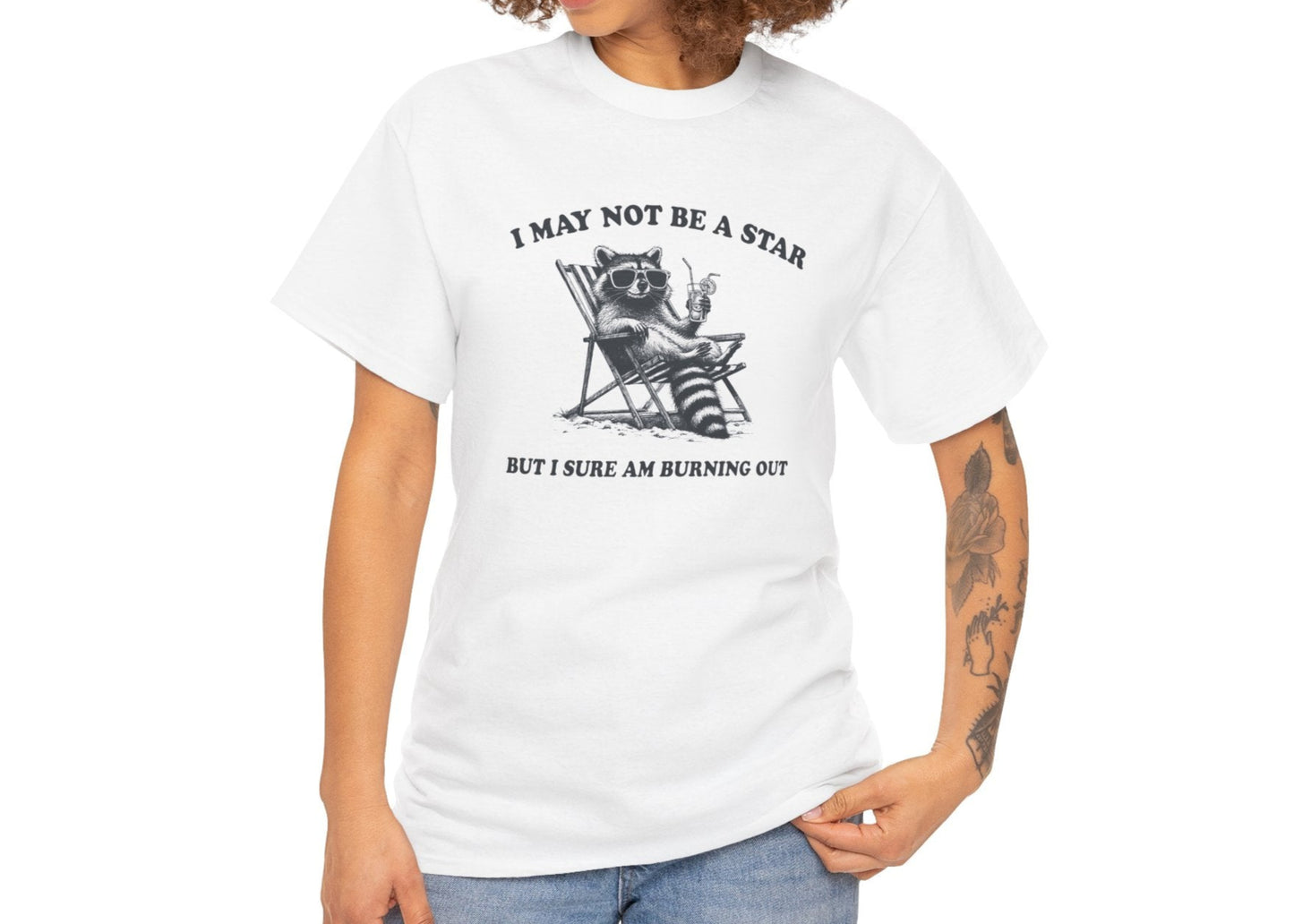 Modeled front view of a white T-shirt featuring a cute raccoon holding a star with text that reads "I May Not Be A Star, But I Sure Am Burning Out." Part of the Vivid Divergence Sensory Friendly Unisex Tees Range.