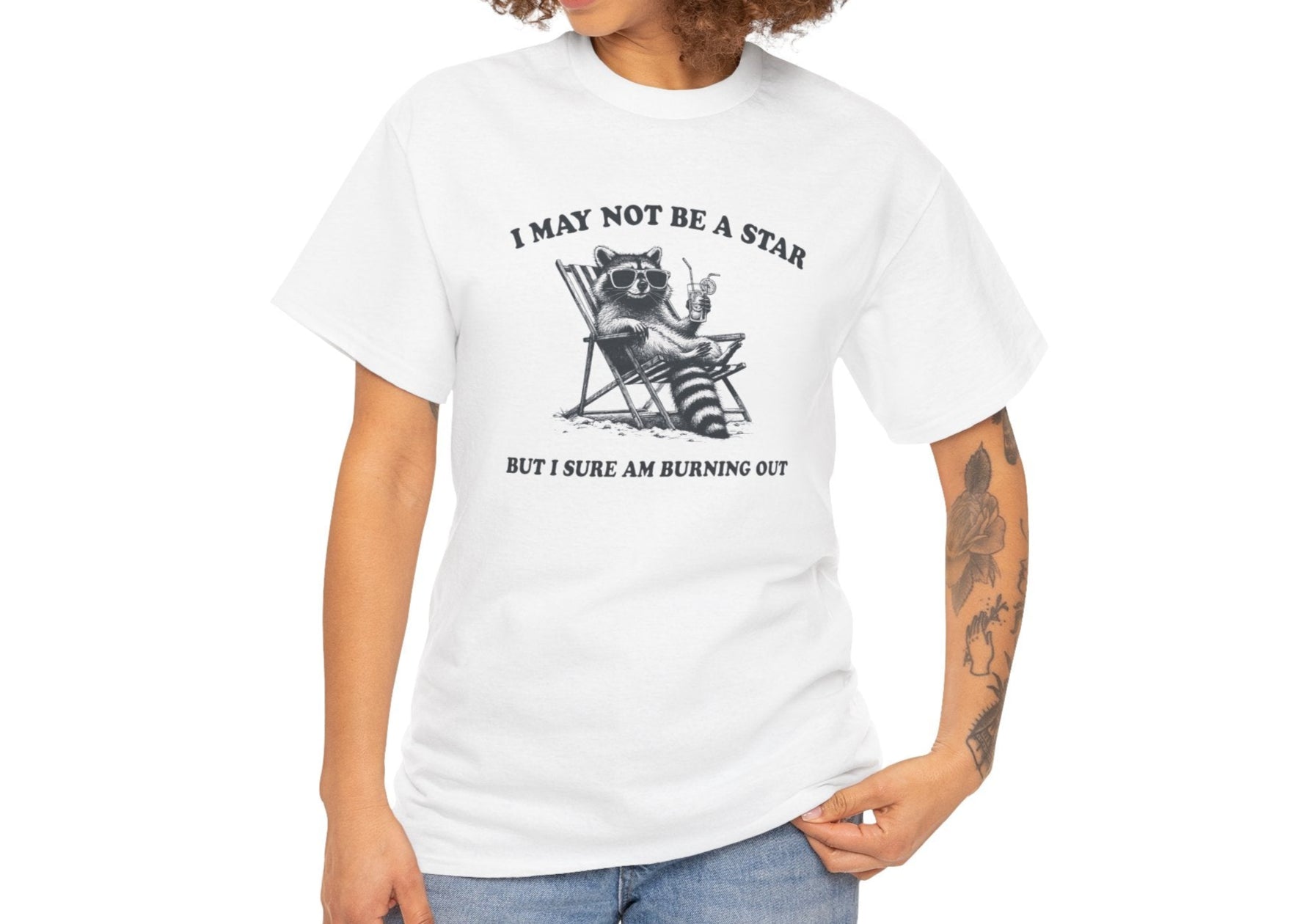 Modeled front view of a white T-shirt featuring a cute raccoon holding a star with text that reads "I May Not Be A Star, But I Sure Am Burning Out." Part of the Vivid Divergence Sensory Friendly Unisex Tees Range.