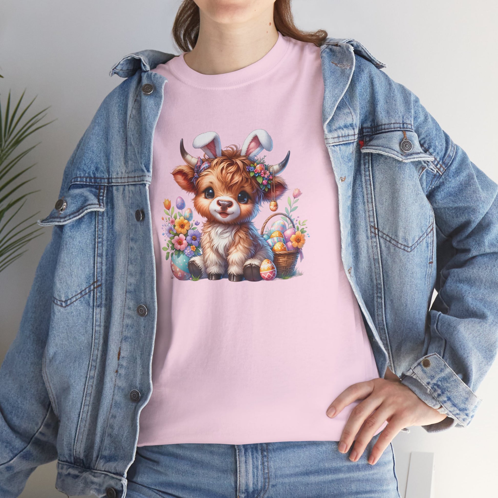 Woman wearing a light pink Easter T-shirt showcasing a fluffy Highland cow with colorful eggs and flowers, a perfect festive choice. Hop into the season in style and shop Easter shirts online today!
